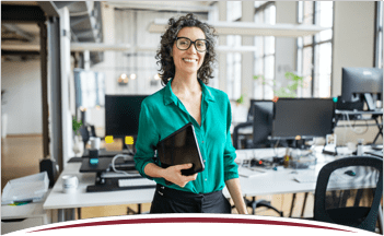 A smiling business manager stands holding a laptop in front of inviting workspaces featuring computers and office ephemera